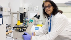 A woman with shoulder length dark hair wearing glasses, a white lab coat and purple latex gloves is leaning over a microscope and looking directly at the camera.
